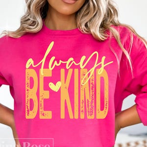 May include: A bright pink sweatshirt with the words "always BE KIND" in yellow script and block letters. A yellow heart is between the words "BE" and "KIND". The sweatshirt is a casual style.