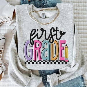 K&ouml;nnte beinhalten: Ein hellgraues Sweatshirt mit den Worten "first grade" in bunten Buchstaben und einer Bleistiftgrafik. Das Sweatshirt wird mit einer blauen Jeans, einer goldenen Halskette und wei&szlig;en Turnschuhen kombiniert.