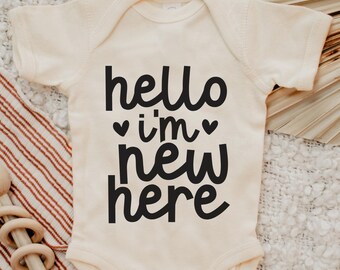 May include: A white baby onesie with black text that reads "hello i'm new here" with two small hearts around the word "i'm".