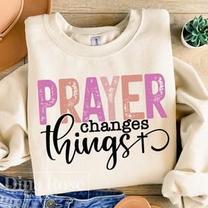 Peut inclure: Un sweat-shirt crème avec le texte "PRAYER changes things" en lettres roses, orange et blanches avec un aspect vieilli. Le texte "things" est en cursive noire avec une croix à la fin.