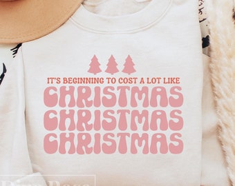 May include: Cream-colored sweatshirt with the text "It's beginning to cost a lot like Christmas" in pink, with three pink Christmas trees above the word "Christmas". A tan hat is visible in the upper left corner.