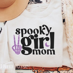 May include: White sweatshirt with black text that reads "spooky girl mom" and a purple skeleton hand making a peace sign.