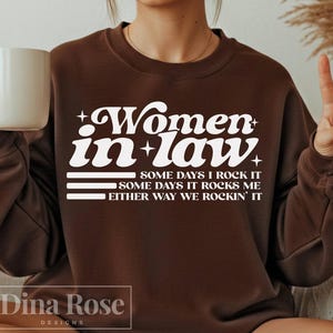 May include: A brown sweatshirt with the text "Women in law" in white, along with the phrases "Some days I rock it", "Some days it rocks me", and "Either way we rockin' it". A white mug is held in the left hand.