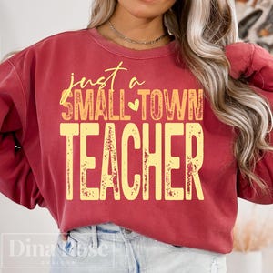 May include: A maroon sweatshirt with the text "just a SMALL TOWN TEACHER" in yellow. The text has a distressed, vintage look. The sweatshirt is a crew neck style.
