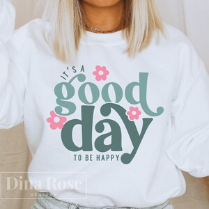Peut inclure: Sweat-shirt blanc avec un motif vert et rose qui dit "C'est une bonne journée pour être heureux" avec des fleurs roses autour du texte.