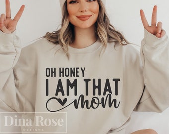Puede incluir: Una sudadera beige con el texto "Oh honey I am that mom" en letras negras con un símbolo de corazón. El texto está centrado en la sudadera. La sudadera la lleva puesta una persona con el pelo rubio y una boina beige.