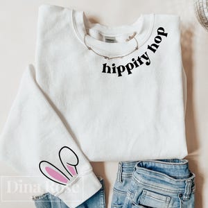 Może przedstawiać: Biała bluza z napisem "hippity hop" czarnymi literami z przodu. Bluza ma różowe uszy królicze na jednym z rękawów.