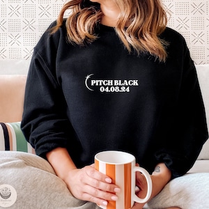Peut inclure: Sweat-shirt noir avec un texte blanc qui dit "Pitch Black 04.08.24". Une personne porte le sweat-shirt et tient une tasse rayée.