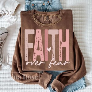 May include: A brown sweatshirt with the words "FAITH over fear" in a distressed pink font. A small white heart is between the words. The sweatshirt is folded on top of blue jeans and a gold chain necklace.