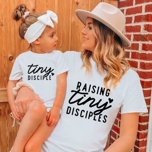 May include: A white t-shirt with the text "Raising tiny disciples" and a small heart. A child is wearing a white t-shirt with the text "tiny disciple" and a small heart.