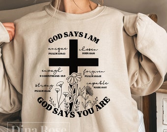 May include: Beige sweatshirt with the text "GOD SAYS I AM" above a black cross and floral design. Below the cross are the words "unique, chosen, enough, forgiven, strong, capable" with corresponding Bible verses. The text "GOD SAYS YOU ARE" is below the floral design.