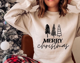 May include: Beige sweatshirt with a black graphic of three Christmas trees and the words "MERRY Christmas" in a script font. The person is wearing red and black plaid pajama pants. A Christmas tree with ornaments is in the background.