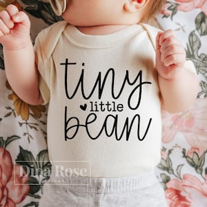 May include: A white baby onesie with black text that reads "tiny little bean" with a small heart symbol before the word "little".