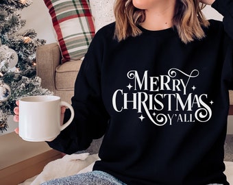 May include: A black sweatshirt with a white graphic that says "Merry Christmas Y'all". The sweatshirt is being worn by a person sitting on a couch with a Christmas tree in the background.