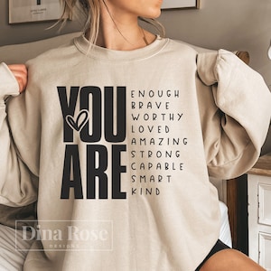 Peut inclure: Un sweat-shirt beige avec les mots "YOU ARE" en grandes lettres noires. Les mots "ENOUGH, BRAVE, WORTHY, LOVED, AMAZING, STRONG, CAPABLE, SMART, KIND" sont écrits en lettres noires plus petites autour des mots "YOU ARE".