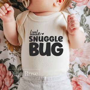 Puede incluir: Body de bebé color crema con la frase "little snuggle bug" en letras negras. El bebé lleva pantalones grises. El fondo tiene un estampado floral. Se ve el nombre de la marca "Dina Rose Designs".