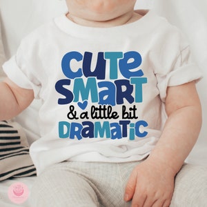 May include: White baby onesie with blue text that reads "Cute Smart & a little bit Dramatic".