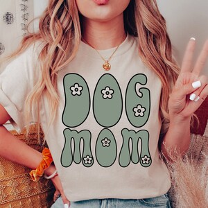 May include: A white t-shirt with a green retro-style graphic that says "Dog Mom" with small white flowers in the letters.