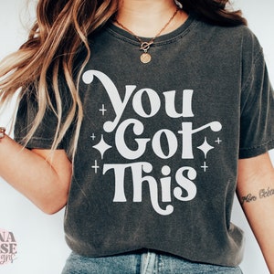 Puede incluir: Una camiseta gris oscuro con el texto "You Got This" en letras blancas. El texto est&aacute; en un estilo de fuente retro con un adorno decorativo. Hay peque&ntilde;as estrellas blancas alrededor del texto.
