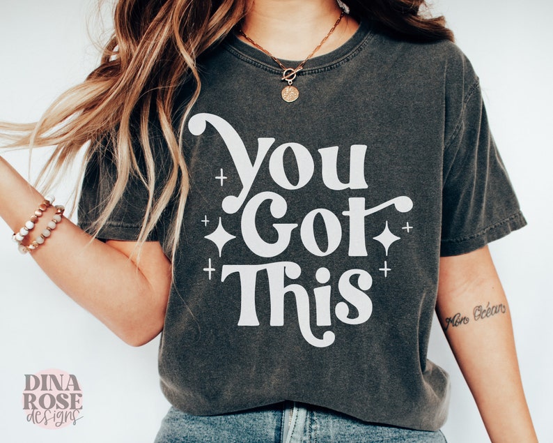 Puede incluir: Una camiseta gris oscuro con el texto "You Got This" en letras blancas. El texto est&aacute; en un estilo de fuente retro con un adorno decorativo. Hay peque&ntilde;as estrellas blancas alrededor del texto.