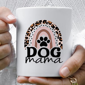 May include: White ceramic mug with a rainbow design featuring a leopard print pattern. The rainbow has a black paw print in the center. The text "DOG mama" is printed below the rainbow.
