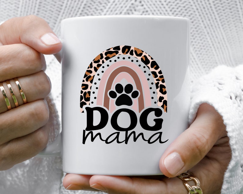 May include: White ceramic mug with a rainbow design featuring a leopard print pattern. The rainbow has a black paw print in the center. The text "DOG mama" is printed below the rainbow.