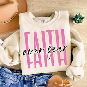 May include: Cream-colored sweatshirt with the words "FAITH OVER FEAR" in pink and black lettering. The sweatshirt is folded on a wooden surface, with a pair of blue jeans and a brown hat in the background.