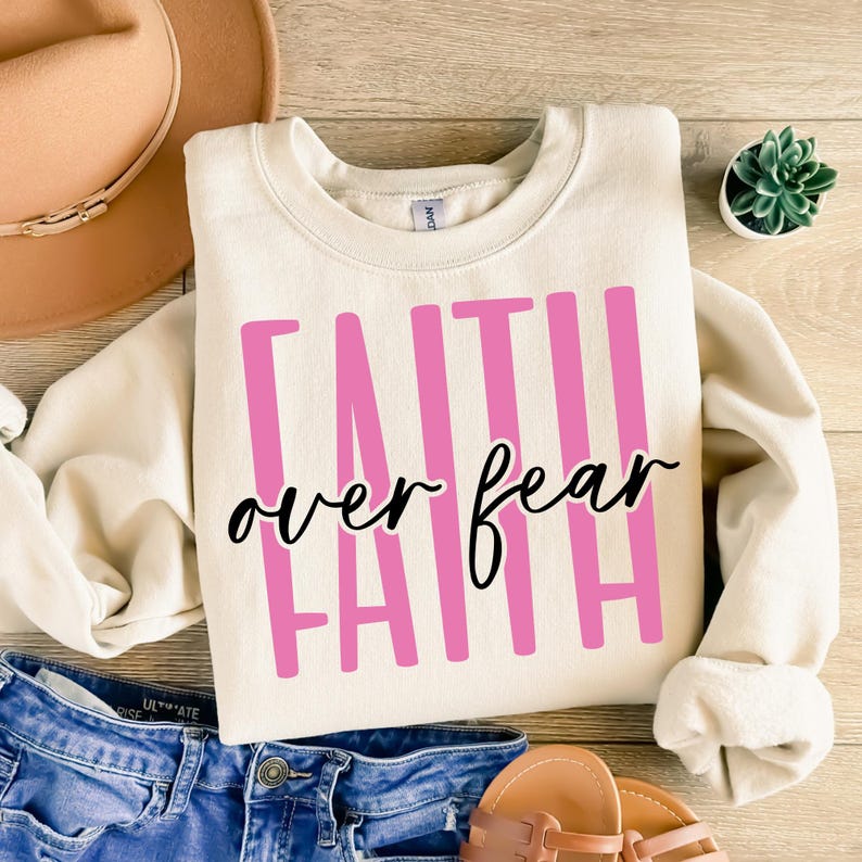 May include: Cream-colored sweatshirt with the words "FAITH OVER FEAR" in pink and black lettering. The sweatshirt is folded on a wooden surface, with a pair of blue jeans and a brown hat in the background.