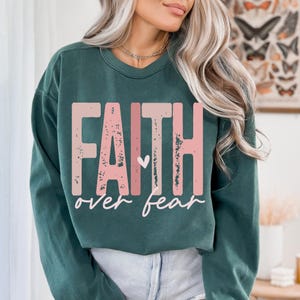 May include: A dark teal sweatshirt with the words "FAITH" in distressed pink block letters above the words "over fear" in a cursive font. A small pink heart is between the two words. The sweatshirt is a solid color.