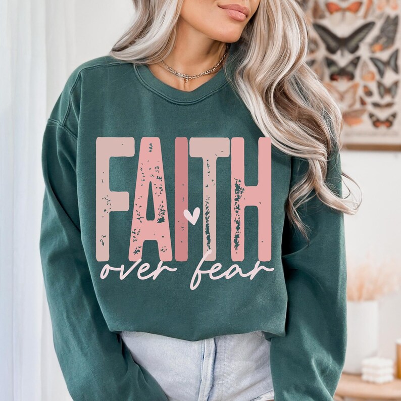 May include: A dark teal sweatshirt with the words "FAITH" in distressed pink block letters above the words "over fear" in a cursive font. A small pink heart is between the two words. The sweatshirt is a solid color.