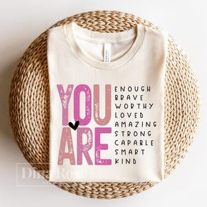 May include: A beige t-shirt with the words "YOU ARE" in pink and white lettering. The words "ENOUGH, BRAVE, WORTHY, LOVED, AMAZING, STRONG, CAPABLE, SMART, KIND" are written in black lettering below the words "YOU ARE".