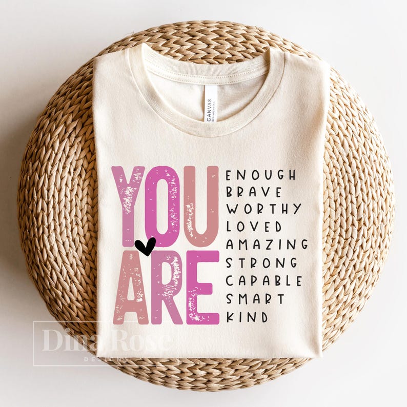 May include: A beige t-shirt with the words "YOU ARE" in pink and white lettering. The words "ENOUGH, BRAVE, WORTHY, LOVED, AMAZING, STRONG, CAPABLE, SMART, KIND" are written in black lettering below the words "YOU ARE".