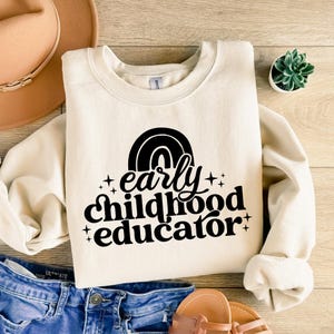 May include: A cream-colored sweatshirt with a black graphic design that says "early childhood educator" with a rainbow above the text.