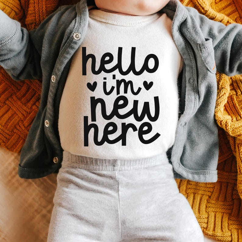May include: A white baby onesie with black text that reads "hello i'm new here" with two small hearts on either side of "i'm".