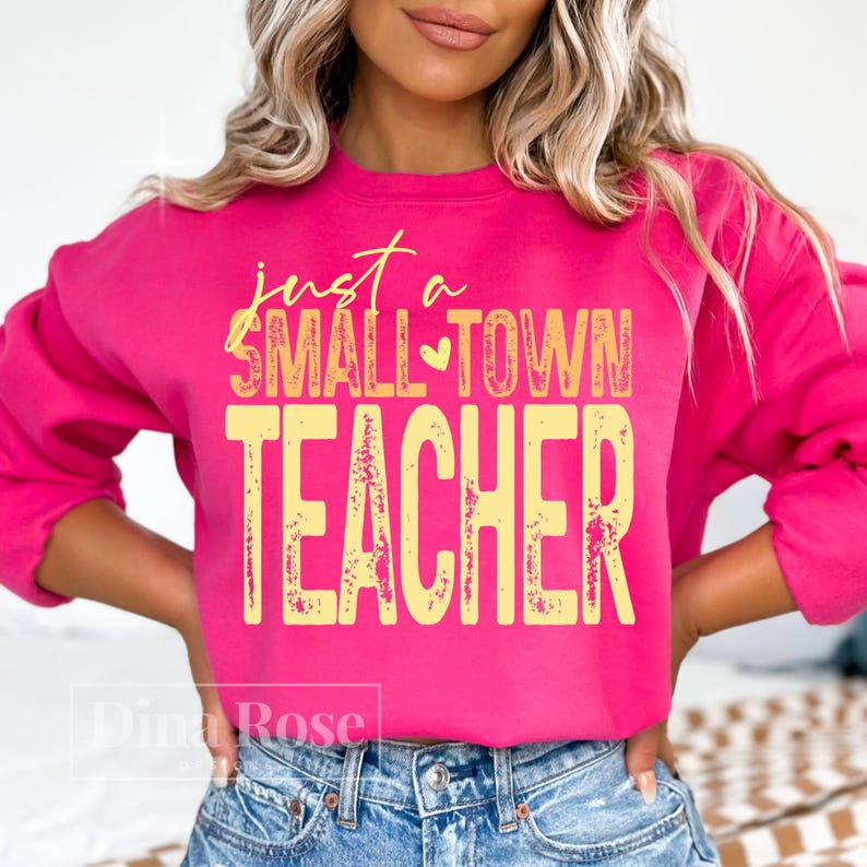 May include: A bright pink sweatshirt with the text "just a SMALL TOWN TEACHER" in yellow. The sweatshirt is a crew neck style and is made of a soft material. The design is centered on the front of the shirt.