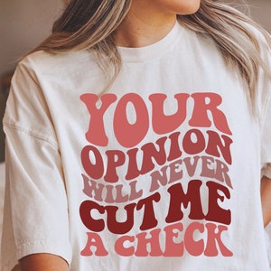 May include: White t-shirt with a red and pink graphic that says "Your opinion will never cut me a check."