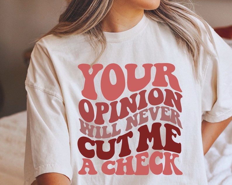 May include: White t-shirt with a red and pink graphic that says "Your opinion will never cut me a check."