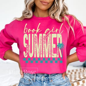 May include: A bright pink sweatshirt with the words "book girl SUMMER" in a light green font. A teal palm tree graphic is on the right side of the word "SUMMER". A teal and white checkered pattern is at the bottom of the word "SUMMER".
