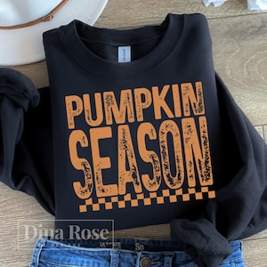 May include: Black sweatshirt with the words "PUMPKIN SEASON" in orange, with a checkered pattern at the bottom. The sweatshirt is folded on top of blue jeans. A white hat is visible in the background.