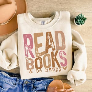 May include: A cream-colored sweatshirt with the text "READ BOOKS & be happy" in pink, brown, and beige letters with a distressed look. A small pink heart is next to the text.