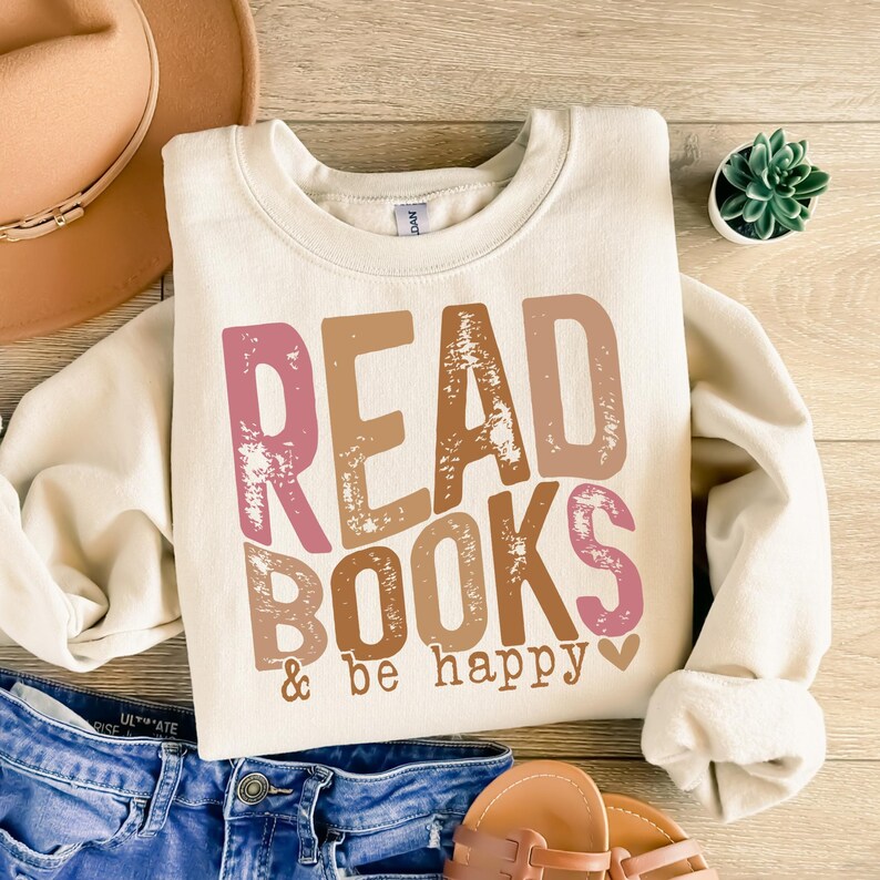 May include: A cream-colored sweatshirt with the text "READ BOOKS & be happy" in pink, brown, and beige letters with a distressed look. A small pink heart is next to the text.