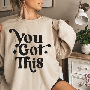 Puede incluir: Una sudadera beige con el texto "You Got This" en letras negras con un estilo de fuente retro. La sudadera tiene cuello redondo y mangas largas.