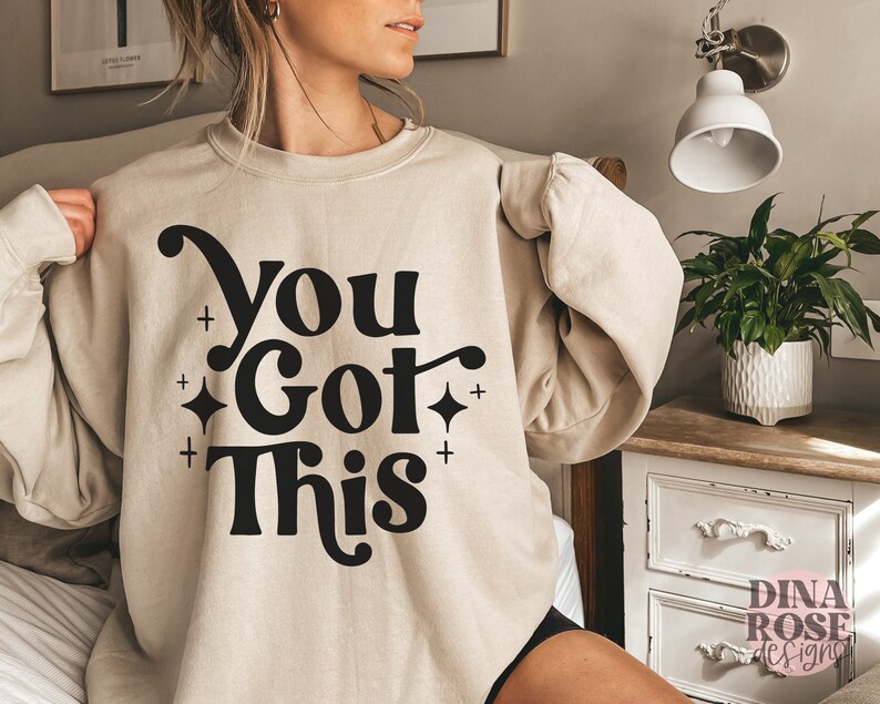 Puede incluir: Una sudadera beige con el texto "You Got This" en letras negras con un estilo de fuente retro. La sudadera tiene cuello redondo y mangas largas.