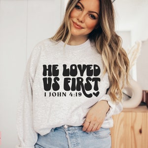 May include: A gray sweatshirt with the text "He Loved Us First 1 John 4:19" in black, retro-style lettering.