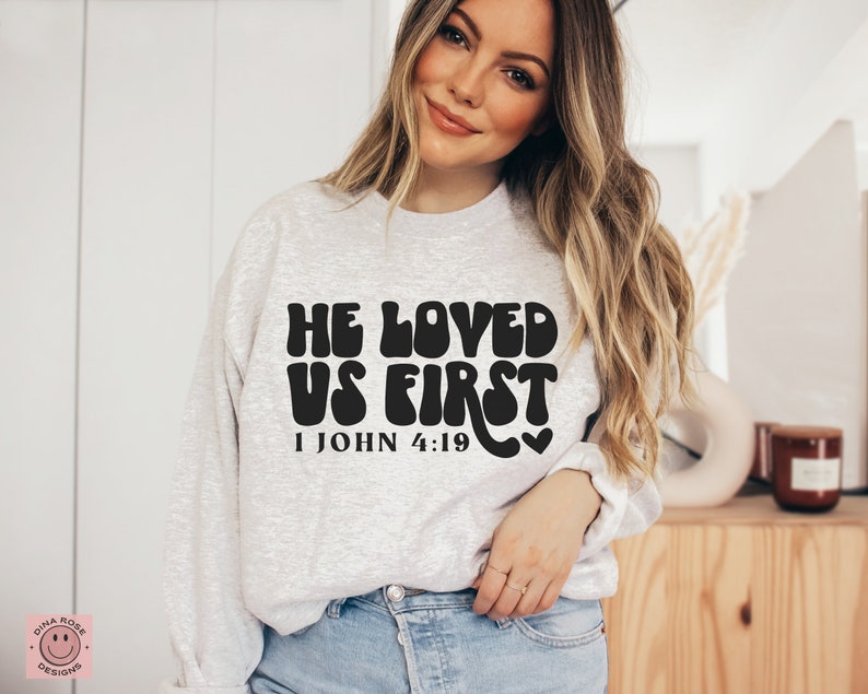 May include: A gray sweatshirt with the text "He Loved Us First 1 John 4:19" in black, retro-style lettering.