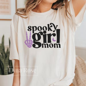 May include: White t-shirt with black text that reads "spooky girl mom" and a purple skeleton hand making a peace sign.