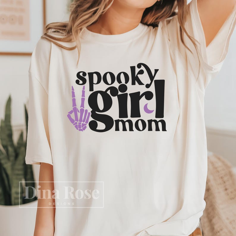 May include: White t-shirt with black text that reads "spooky girl mom" and a purple skeleton hand making a peace sign.