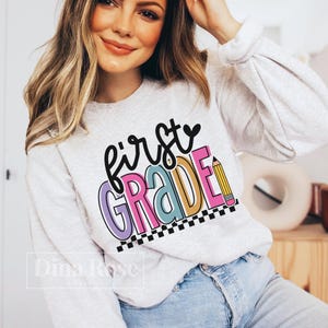 K&ouml;nnte beinhalten: Ein hellgraues Sweatshirt mit dem Schriftzug "first GRADE" in farbenfrohen, verspielten Buchstaben. Das Design beinhaltet eine Bleistiftgrafik und einen schwarz-wei&szlig;en Schachbrettmuster-Rand, ideal f&uuml;r den Schulanfang. Das Sweatshirt ist ein bequemer, l&auml;ssiger Stil.