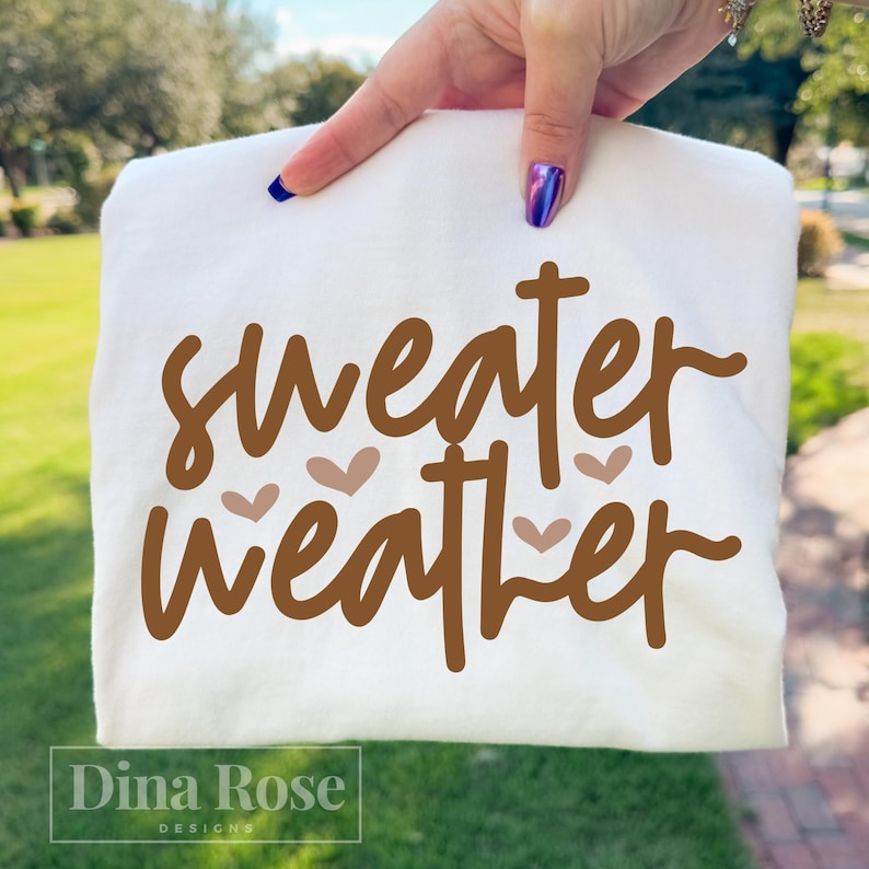 May include: A white sweatshirt with the words "sweater weather" in brown script, accented with small heart shapes. The Dina Rose Designs logo is at the bottom.