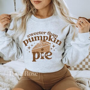 Pode incluir: Uma camisa de moletom cinza com o texto "sweeter than pumpkin pie" em letras marrons. Uma imagem estilizada de uma fatia de torta de ab&oacute;bora est&aacute; no centro do texto.